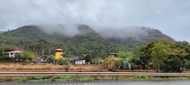 A scenic landscape with mist-covered hills in the background, dotted with lush green trees and scattered houses with colorful roofs. A prominent highway runs in the foreground, while the overall setting appears serene and calm.