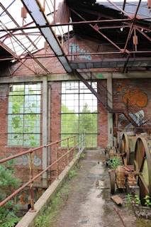 An abandoned industrial structure with rusted metal beams and broken windows. Overgrown plants are visible through the windows, indicating nature reclaiming the space. Graffiti adorns the brick walls, and old machinery parts are scattered on the ground. The overall atmosphere suggests neglect and decay.