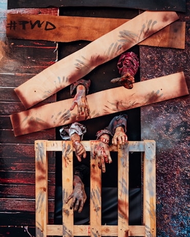 Several creepy hands with a zombie-like appearance protrude through wooden boards and a wooden pallet. The boards have what seems to be painted handprints, and the overall setting looks worn and eerie.