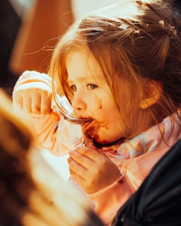 A young child is eating with a spoon, getting food smeared around their mouth. The child has light brown hair and is wearing a pink outfit. Sunlight casts a warm glow on their face, highlighting their focused expression.