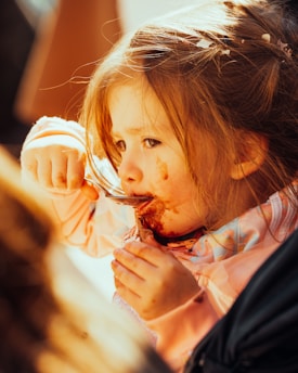 A young child is eating with a spoon, getting food smeared around their mouth. The child has light brown hair and is wearing a pink outfit. Sunlight casts a warm glow on their face, highlighting their focused expression.