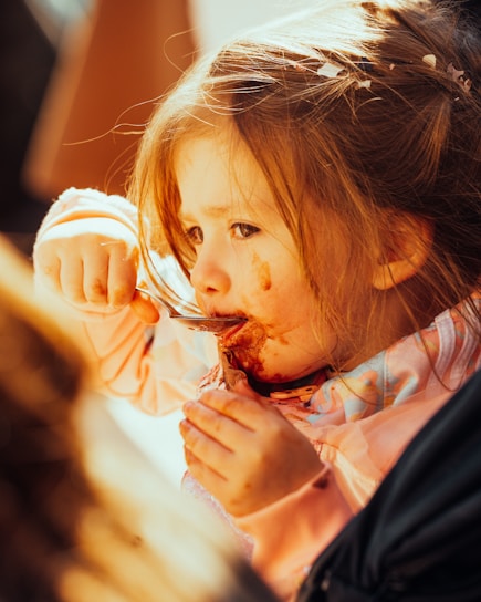 A young child is eating with a spoon, getting food smeared around their mouth. The child has light brown hair and is wearing a pink outfit. Sunlight casts a warm glow on their face, highlighting their focused expression.