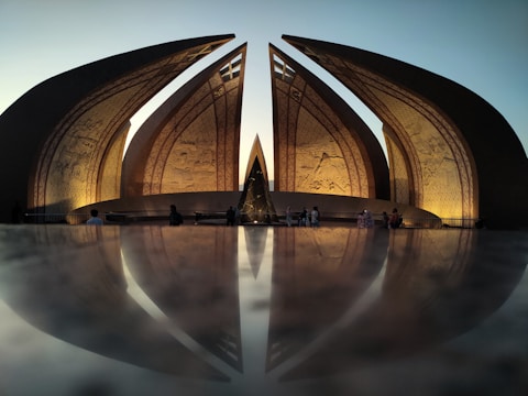 A monumental structure with large petal-shaped granite slabs that are artfully illuminated. The slabs have intricate carvings, and their reflection can be seen on a polished surface below, creating a symmetrical visual effect. Several people are visible, walking or sitting near the monument, adding to the scene's scale and grandeur.