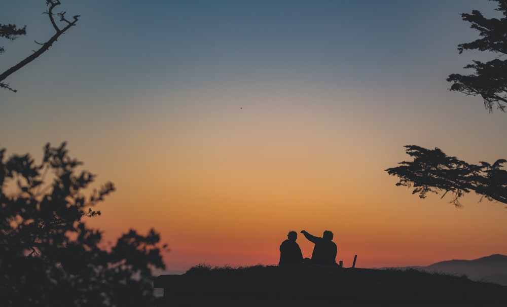 a silhouette of a couple of people sitting on a hill at sunset