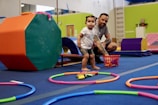 A young child is standing inside a play area with colorful gym equipment and toys scattered on the floor. An adult is crouching nearby, observing or interacting with the child. The surroundings are brightly colored with various play and exercise items.