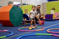 A young child is standing inside a play area with colorful gym equipment and toys scattered on the floor. An adult is crouching nearby, observing or interacting with the child. The surroundings are brightly colored with various play and exercise items.
