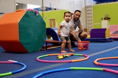 A young child is standing inside a play area with colorful gym equipment and toys scattered on the floor. An adult is crouching nearby, observing or interacting with the child. The surroundings are brightly colored with various play and exercise items.
