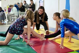 Four girls are practicing gymnastics on colorful mats in a gymnasium setting. They are engaged in stretching exercises, each wearing different gymnastic attire. A woman, possibly a coach, kneels in the center and appears to be giving instructions. In the background, there are several people watching, some seated and some standing.