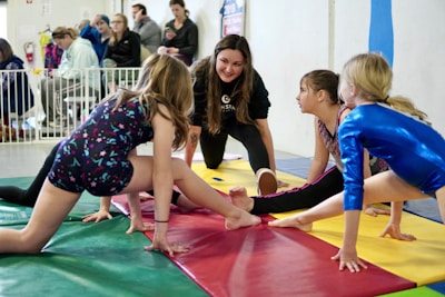 Four girls are practicing gymnastics on colorful mats in a gymnasium setting. They are engaged in stretching exercises, each wearing different gymnastic attire. A woman, possibly a coach, kneels in the center and appears to be giving instructions. In the background, there are several people watching, some seated and some standing.