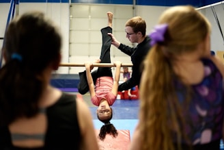 A young girl in a pink shirt is hanging upside down from a gymnastics bar, with a coach assisting her. In the foreground, there are two other children watching, one with a long ponytail and a purple bow. The scene takes place in a gymnasium with mats on the floor and equipment around.
