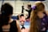 A young girl in a pink shirt is hanging upside down from a gymnastics bar, with a coach assisting her. In the foreground, there are two other children watching, one with a long ponytail and a purple bow. The scene takes place in a gymnasium with mats on the floor and equipment around.
