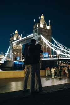A nighttime scene featuring a couple embracing near an illuminated bridge with distinctive architecture. The bridge's lights create reflections on the water and emphasize its structure. Several other people are visible in the background, casually walking and socializing. The cityscape behind provides an urban ambiance.