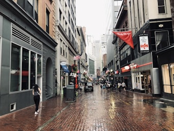 A city street scene featuring wet brick pavement, several people walking, and buildings with storefronts. American flags are hung along the street, and a red sign prominently displays 'Tasty Burger.' Tall buildings surround the narrow street, creating an urban atmosphere.