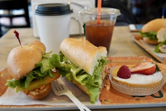 A neatly arranged sandwich filled with fresh lettuce and tomatoes is served on a tray next to a small dessert topped with fresh strawberries and a raspberry. In the background, a cup of coffee with a lid and a plastic cup filled with iced tea or coffee accompanied by a straw are visible. A fork is placed in front of the sandwich on the tray.