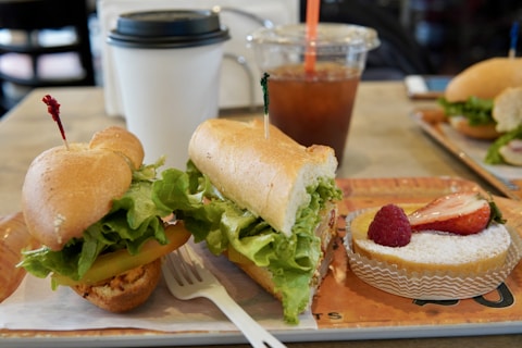 A neatly arranged sandwich filled with fresh lettuce and tomatoes is served on a tray next to a small dessert topped with fresh strawberries and a raspberry. In the background, a cup of coffee with a lid and a plastic cup filled with iced tea or coffee accompanied by a straw are visible. A fork is placed in front of the sandwich on the tray.