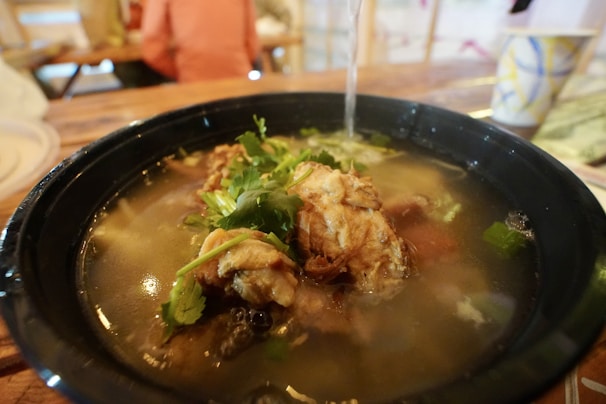 A steaming bowl of soup filled with tender pieces of chicken, fresh cilantro, and vegetables in a rich, clear broth. The bowl is placed on a wooden table, with a softly blurred background suggesting a casual dining setting.