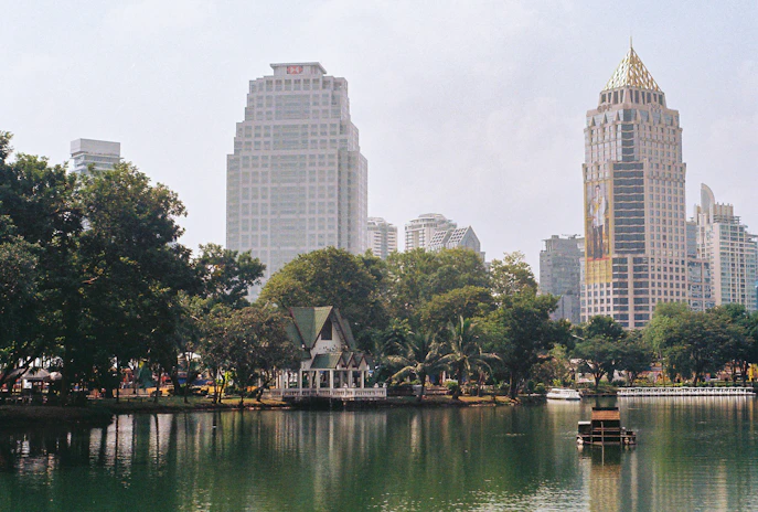 a body of water with trees and buildings in the background with Lumphini Park in the background