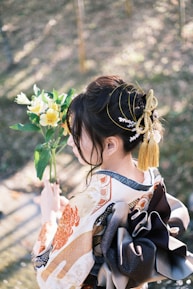 A person dressed in a traditional, elaborately designed kimono with intricate patterns and a large, ornate obi tied in the back. The person is holding a bouquet of white and yellow flowers, and their hair is styled elegantly with decorative accessories, including a tassel and delicate adornments. Soft, dappled sunlight filters through trees in the background, creating a serene garden setting.