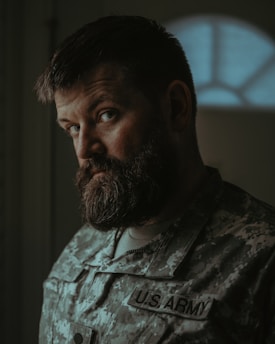A man with a thick beard and a serious expression wearing a U.S. Army camouflage uniform is standing indoors. The lighting is dim, creating a shadowed and introspective atmosphere. The focus is on the man's face, while the background is blurred.