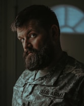 A man with a thick beard and a serious expression wearing a U.S. Army camouflage uniform is standing indoors. The lighting is dim, creating a shadowed and introspective atmosphere. The focus is on the man's face, while the background is blurred.