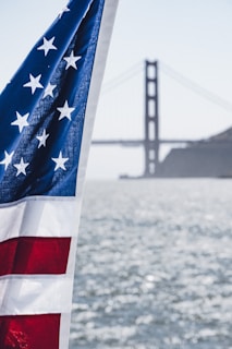 An American flag flutters prominently in the foreground with a blurred view of the Golden Gate Bridge in the background across a body of water. The flag displays stars and stripes in red, white, and blue, partially illuminated by sunlight. The scene conveys a sense of patriotism and iconic American imagery.