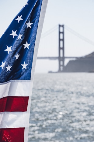 An American flag flutters prominently in the foreground with a blurred view of the Golden Gate Bridge in the background across a body of water. The flag displays stars and stripes in red, white, and blue, partially illuminated by sunlight. The scene conveys a sense of patriotism and iconic American imagery.