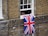 A brick building with a partially open window features a Union Jack flag hanging out. Part of a person's body is visible through the window. The bricks are various shades of brown and gray, and a security camera is mounted below the window.