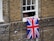 A brick building with a partially open window features a Union Jack flag hanging out. Part of a person's body is visible through the window. The bricks are various shades of brown and gray, and a security camera is mounted below the window.