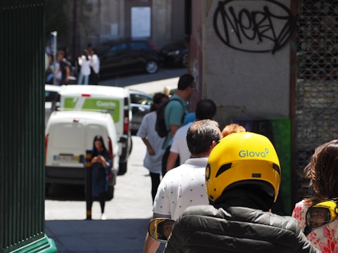 A group of people are walking on a street, with one individual wearing a bright yellow Glovo helmet. In the background, there is a white van with a logo, a woman using a smartphone, and graffiti on a nearby wall.
