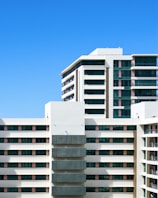 A modern residential building with multiple floors and large windows, featuring a clean architectural design. The facade is mainly white with accents of dark glass panels and some wooden elements. The sky in the background is clear and blue.