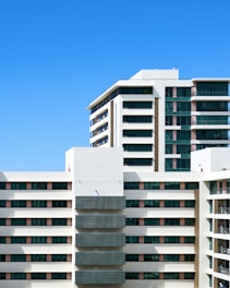 A modern residential building with multiple floors and large windows, featuring a clean architectural design. The facade is mainly white with accents of dark glass panels and some wooden elements. The sky in the background is clear and blue.