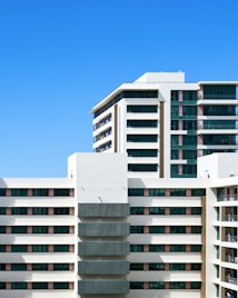 A modern residential building with multiple floors and large windows, featuring a clean architectural design. The facade is mainly white with accents of dark glass panels and some wooden elements. The sky in the background is clear and blue.