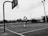A black and white photograph of an outdoor basketball court with a person in mid-air, shooting a basketball towards the hoop. The court appears empty, surrounded by fencing and some overhead lights. The background shows a few buildings and some trees.