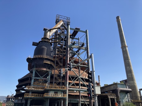 A large industrial structure with complex metal framework and towering smokestacks reaches into a clear blue sky. The facility includes prominent cylindrical components and various pipelines. A traffic light is visible in the foreground, adding to the urban industrial scene.