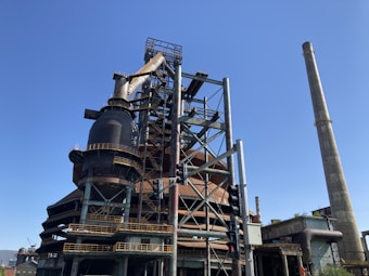 A large industrial structure with complex metal framework and towering smokestacks reaches into a clear blue sky. The facility includes prominent cylindrical components and various pipelines. A traffic light is visible in the foreground, adding to the urban industrial scene.