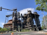 A large industrial complex with massive metal structures and intricate pipework, set under a clear blue sky. Trees and a traffic light are visible in the foreground, while the complex towers overhead.