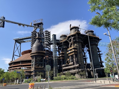 A large industrial complex with massive metal structures and intricate pipework, set under a clear blue sky. Trees and a traffic light are visible in the foreground, while the complex towers overhead.
