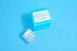 A jar and a box of skincare product labeled 'Dermatology' are placed against a solid light blue background. The jar appears to be small with a white lid and blue-green label, while the accompanying box is larger with similar labeling and a bold cyan color.
