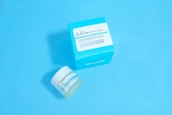 A jar and a box of skincare product labeled 'Dermatology' are placed against a solid light blue background. The jar appears to be small with a white lid and blue-green label, while the accompanying box is larger with similar labeling and a bold cyan color.