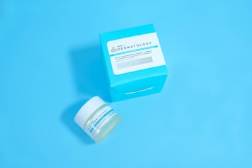 A jar and a box of skincare product labeled 'Dermatology' are placed against a solid light blue background. The jar appears to be small with a white lid and blue-green label, while the accompanying box is larger with similar labeling and a bold cyan color.