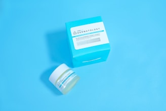 A jar and a box of skincare product labeled 'Dermatology' are placed against a solid light blue background. The jar appears to be small with a white lid and blue-green label, while the accompanying box is larger with similar labeling and a bold cyan color.