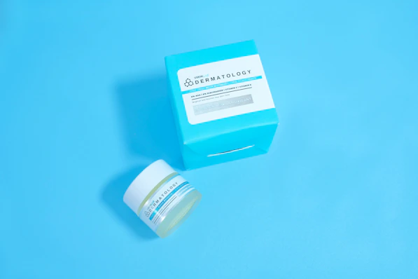 A jar and a box of skincare product labeled 'Dermatology' are placed against a solid light blue background. The jar appears to be small with a white lid and blue-green label, while the accompanying box is larger with similar labeling and a bold cyan color.