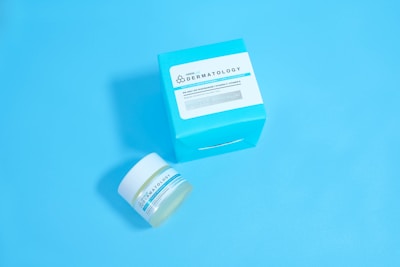 A jar and a box of skincare product labeled 'Dermatology' are placed against a solid light blue background. The jar appears to be small with a white lid and blue-green label, while the accompanying box is larger with similar labeling and a bold cyan color.