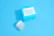 A jar and a box of skincare product labeled 'Dermatology' are placed against a solid light blue background. The jar appears to be small with a white lid and blue-green label, while the accompanying box is larger with similar labeling and a bold cyan color.
