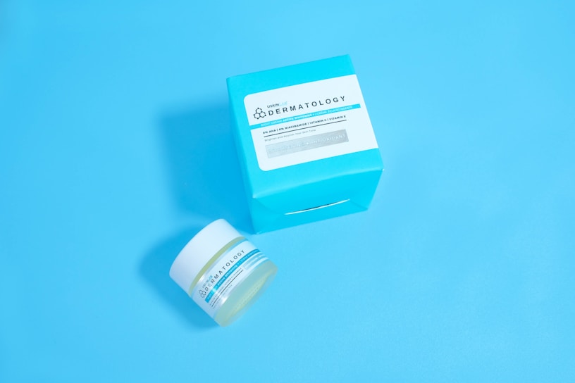 A jar and a box of skincare product labeled 'Dermatology' are placed against a solid light blue background. The jar appears to be small with a white lid and blue-green label, while the accompanying box is larger with similar labeling and a bold cyan color.