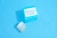 A jar and a box of skincare product labeled 'Dermatology' are placed against a solid light blue background. The jar appears to be small with a white lid and blue-green label, while the accompanying box is larger with similar labeling and a bold cyan color.