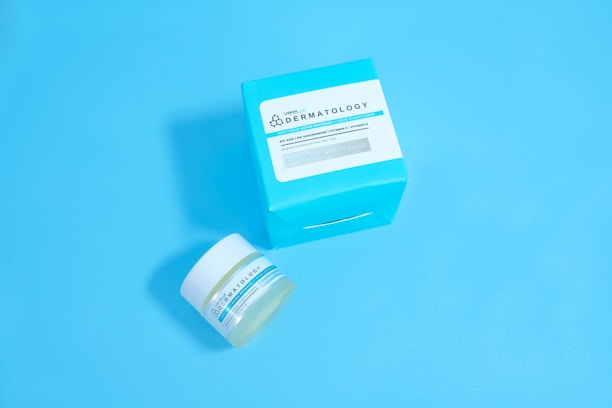 A jar and a box of skincare product labeled 'Dermatology' are placed against a solid light blue background. The jar appears to be small with a white lid and blue-green label, while the accompanying box is larger with similar labeling and a bold cyan color.