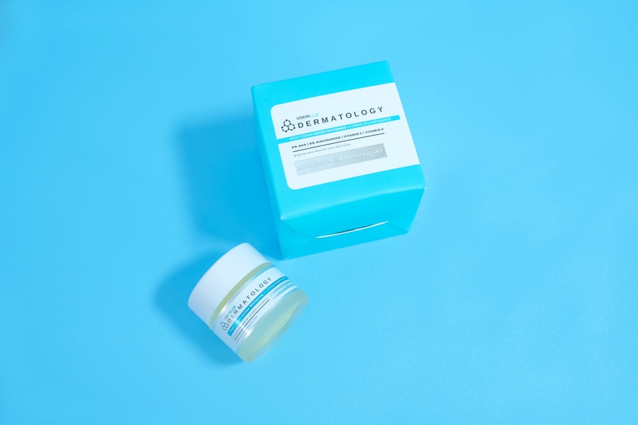 A jar and a box of skincare product labeled 'Dermatology' are placed against a solid light blue background. The jar appears to be small with a white lid and blue-green label, while the accompanying box is larger with similar labeling and a bold cyan color.