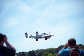 A vintage military aircraft is flying low in the sky with two spectators in the foreground capturing the moment with cameras. The plane has distinctive red and white stripes on its tail fins. Below, there's a backdrop of green trees contrasting with the clear blue sky.