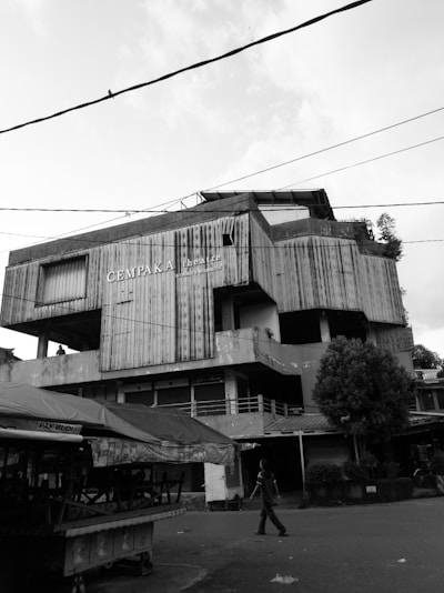A black and white photograph features a large, worn-down building with several stories. The structure is marked by corrugated metal panels and a sign that reads 'CemPAKA Theatre' on its facade. The foreground includes empty market stalls with tarps and a solitary person walking past the building. Overhead, electrical wires crisscross the sky, and a bird is perched on one of the wires.